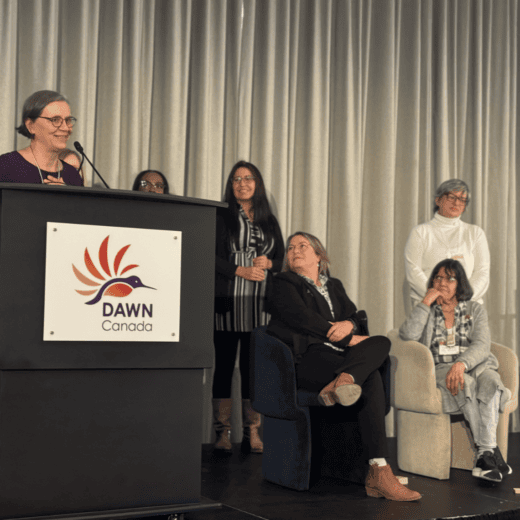 Photo from the 2023 Hummingbird Awards ceremony in March 2024. Deborah Stienstra is standing behind a podium and talking in a microphone, while other attendees are standing on stage listening and looking at them.