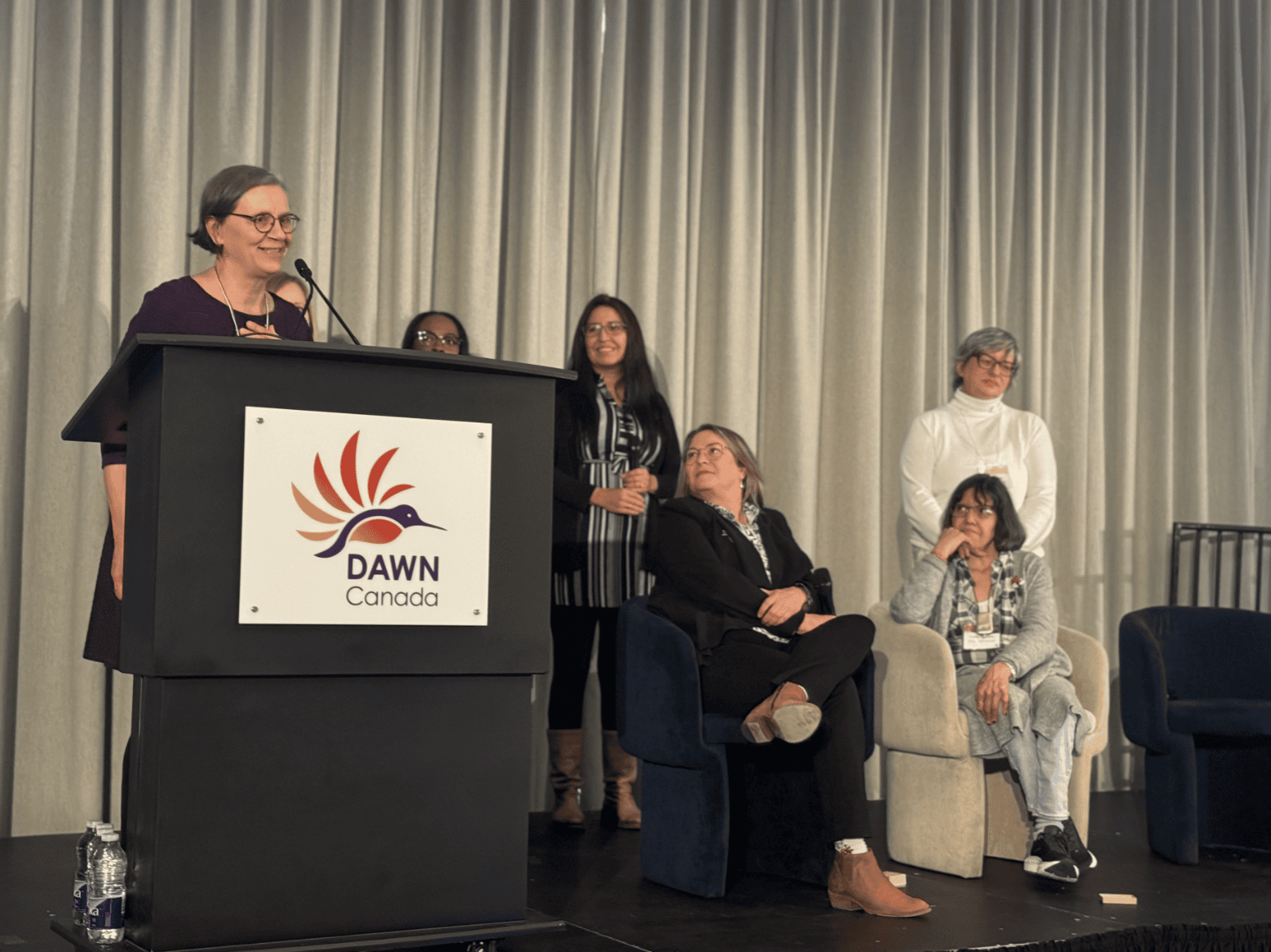 Photo from the 2023 Hummingbird Awards ceremony in March 2024. Deborah Stienstra is standing behind a podium and talking in a microphone, while other attendees are standing on stage listening and looking at them.