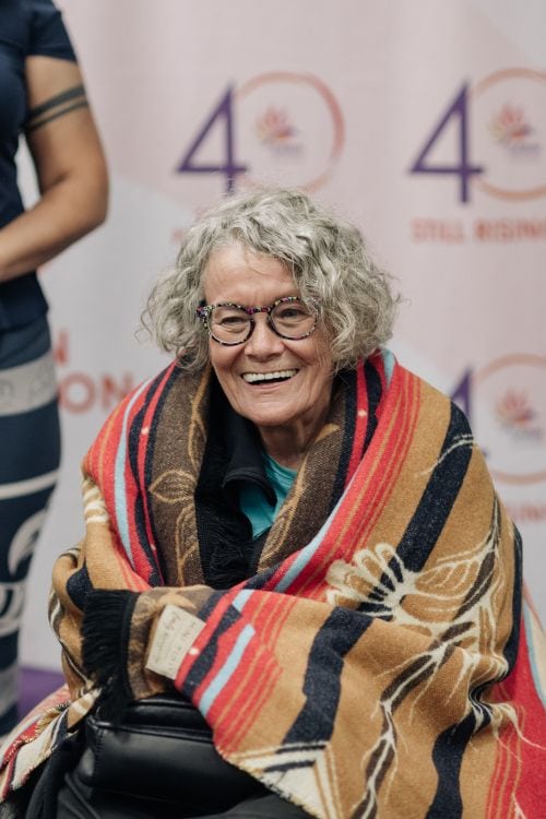 BILINGUAL EN/FR. EN: This is an image of Pat Danforth seated in her wheelchair, draped in a colourful patterned blanket. She smiles warmly, wearing glasses, with curly grey hair. Behind her is a backdrop featuring DAWN Canada’s “40 Years – Still Rising!” anniversary logo. The image conveys a joyful and celebratory moment. FR: Ceci est une image de Pat Danforth assise dans son fauteuil roulant, enveloppée dans une couverture colorée à motifs. Elle sourit chaleureusement, porte des lunettes et a les cheveux gris et bouclés. En arrière-plan, on aperçoit une bannière avec le logo du 40e anniversaire de DAWN Canada « 40 ans – Toujours en pleine ascension ! ». L’image dégage une atmosphère joyeuse et festive.