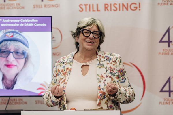 Bonnie Brayton, CEO of DAWN Canada, speaks at a podium during the organization’s 40th Anniversary celebration. She is wearing a floral blazer and glasses, smiling as she addresses the audience. Behind her is a banner featuring DAWN’s “40 Years – Still Rising!” anniversary branding, and a screen displaying an image of the presenter. The setting is bright and celebratory.