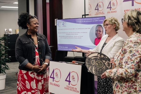 BILINGUAL EN/FR EN: Three individuals are gathered at DAWN Canada’s 40th Anniversary celebration. One person stands on the left, smiling and facing the two others, who are positioned near a podium and holding a circular award plaque. Behind them is a screen displaying event details and a photo of a woman being honoured, along with a backdrop featuring DAWN Canada’s “40 Years – Still Rising!” logo in English and French. The moment appears warm and respectful. FR: Trois personnes se tiennent ensemble devant une bannière de fond officielle lors de la célébration du 40e anniversaire de DAWN Canada. Une personne est debout à un podium, souriante, tandis qu’une deuxième personne à ses côtés tient une plaque circulaire représentant un prix. La troisième personne, vêtue d’une robe sans manches, sourit et fait un geste avec enthousiasme. Derrière elles se trouve un mur médiatique avec le logo du 40e anniversaire de DAWN Canada « 40 ans – Toujours en pleine ascension ! » en anglais et en français. L’atmosphère est joyeuse et festive.