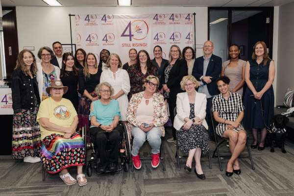 A group photo taken at DAWN Canada’s 40th Anniversary celebration. The image shows 21 individuals of diverse ages, races, and genders. Five people are seated at the front, while the remaining individuals stand behind them. They are gathered in front of a step-and-repeat banner featuring DAWN’s 40th anniversary logo in both English and French. The setting is a well-lit office space with a warm, celebratory atmosphere. A guide dog is visible on the far right.