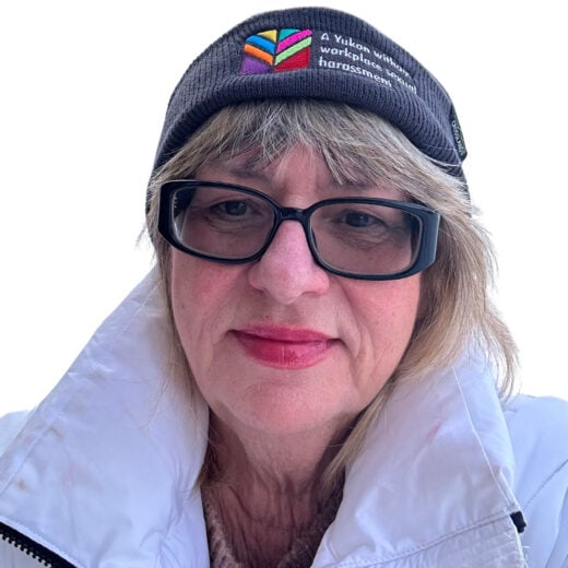 A woman wearing glasses and a dark beanie that reads 'A Yukon without workplace sexual harassment.' She has shoulder-length blonde hair, red lipstick, and is dressed in a white winter jacket. The background is white
