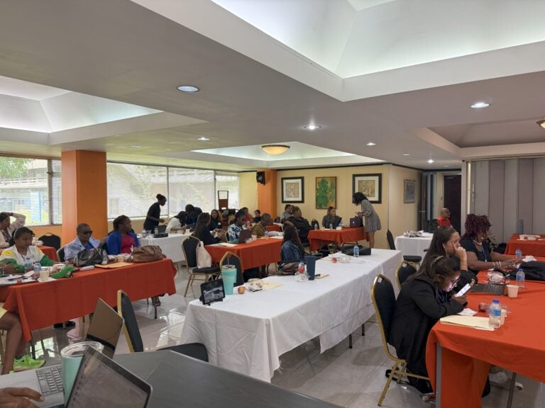 A group of participants seated at tables covered with orange and white tablecloths, engaging in discussion and note-taking during a symposium in Trinidad and Tobago. Some people are writing, using laptops, or talking in small groups in a bright room with large windows and framed artwork on the walls | Un groupe de participant·e·s assis à des tables couvertes de nappes orange et blanches, prenant part à des discussions et prenant des notes lors d’un symposium à Trinité-et-Tobago. Certaines personnes écrivent, utilisent des ordinateurs portables ou discutent en petits groupes dans une salle lumineuse avec de grandes fenêtres et des œuvres encadrées sur les murs.