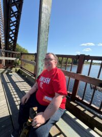 a Non-Binary person, smiling wearing bright tshirt, holding a water bottle in their left hand, sitting on a bench outside with a body of water behind them