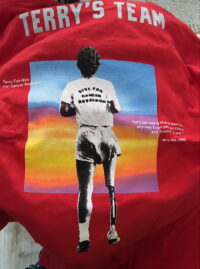 close up of red tshirt with graphic white writing that says "Terry's Team" and an illustrated image of the back of Terry Fox doing his marathon, set to a multitone sunset square with writing in top left and middle right side of the sunset square. Top left writing says: " 'Terry Fox Run for Cancer Research" and middle right says: "All I can say is that if there is any way I can get out there and finish it, I will.' Terry Fox, 1980"