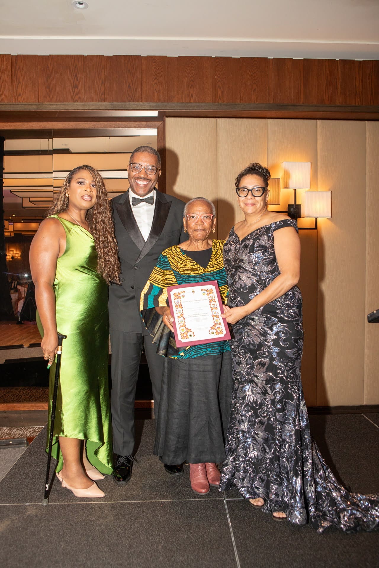 Four people pose together at a gala, smiling as an older woman holds a framed certificate, surrounded by three elegantly dressed attendees.